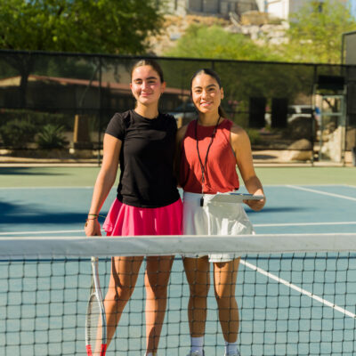 Cheerful caucasian teen girl and latin woman coach on the tennis court smiling while at tennis school