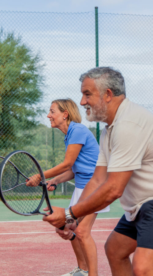 senior tennis couple is captured from the side, holding their rackets and preparing to play during a match on the court. Their focused expressions show their readiness and enthusiasm for the game